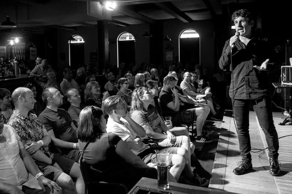 A stand-up comedian performing to an audience at The Charles Bradlaugh